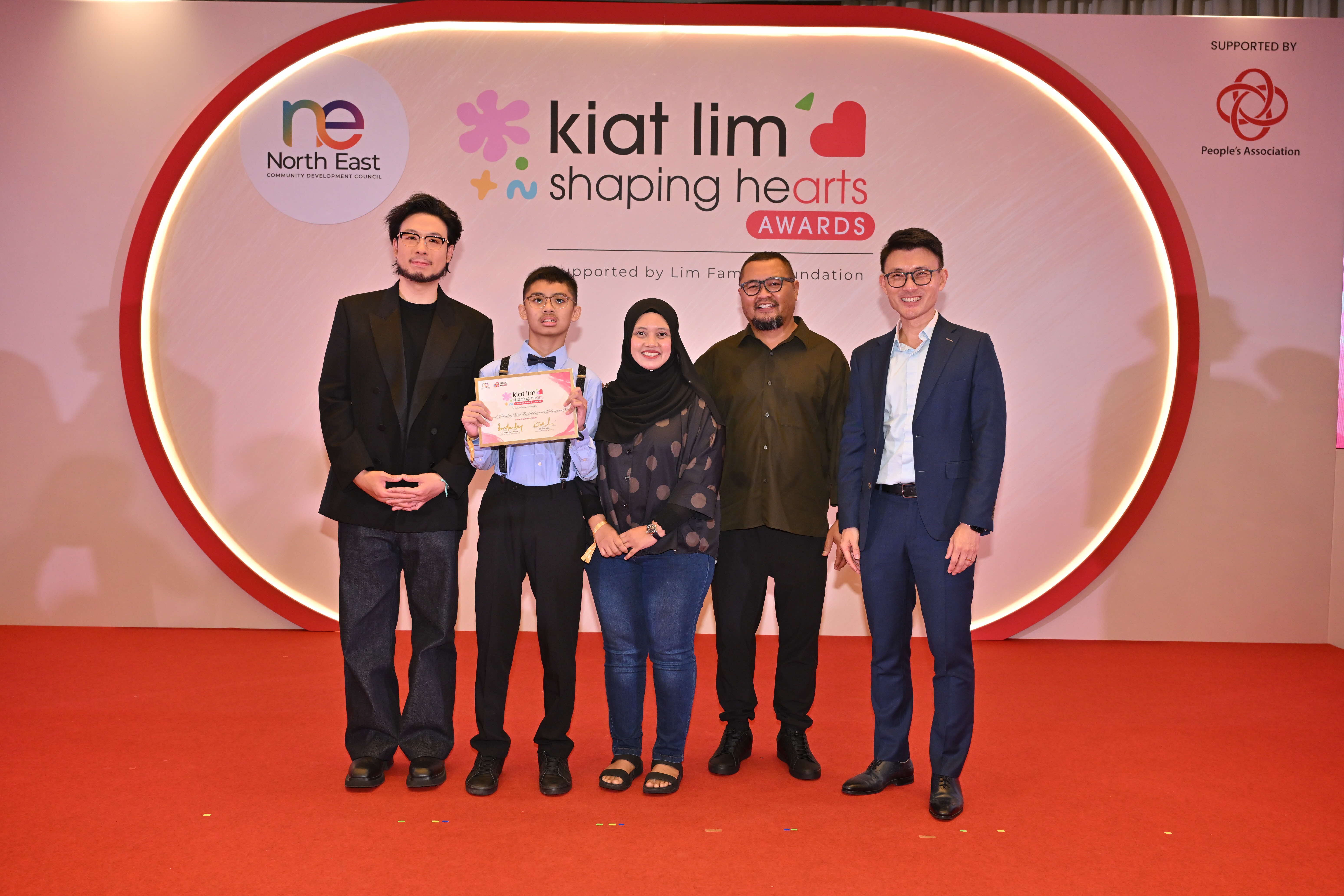 Award recipient standing on stage with the presenter, holding their certificate and posing for a group photo during the award ceremony, with a backdrop reading ‘Kiat Lim-Shaping Hearts Awards'.